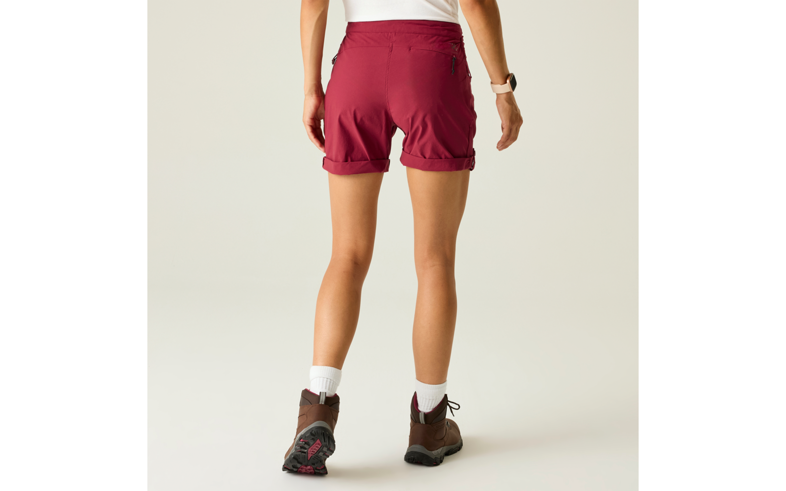 Rückansicht einer stehenden Person in bordeauxroten Shorts, weißen Socken und braunen Wanderschuhen vor neutralem Hintergrund