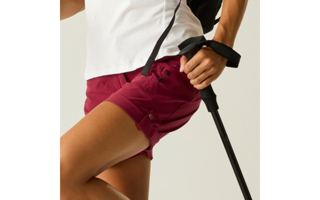 Teilansicht einer Person mit weißem Oberteil und bordeauxroten Shorts, die einen schwarzen Trekkingstock hält; schwarzer Rucksackriemen sichtbar