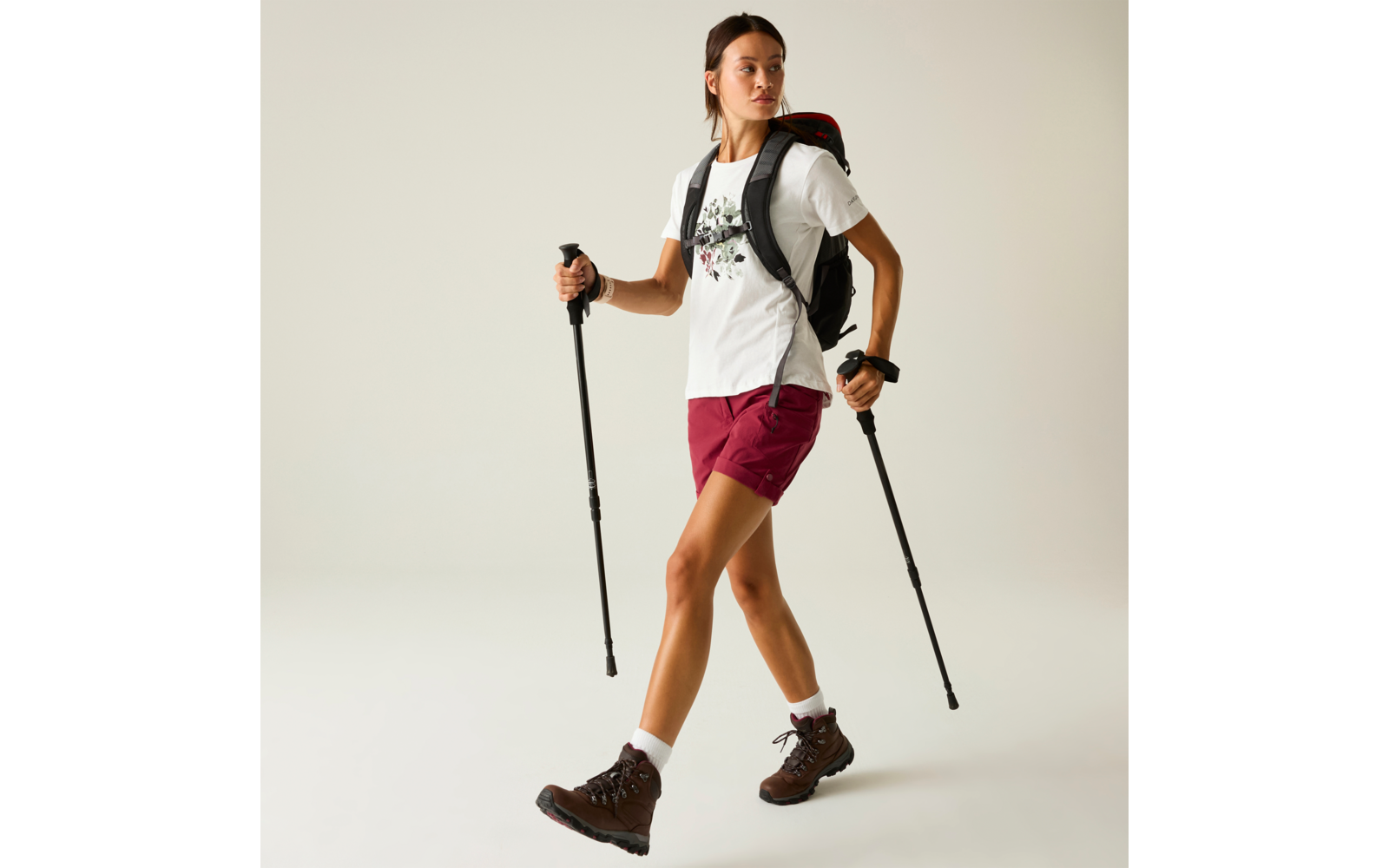 Person mit Zopf trägt weißes T‑Shirt mit Blumenmotiv, bordeauxrote Shorts, braune Wanderschuhe, weiße Socken, schwarzer Rucksack mit roter Innenfarbe; hält zwei schwarze Trekkingstöcke; heller einfarbiger Hintergrund