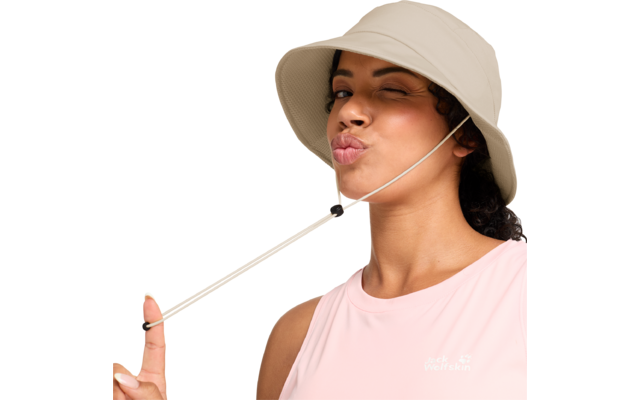 Person mit beigem Anglerhut zwinkert, macht einen Kussmund, hält den Hutriemen und trägt ein hellrosa ärmelloses Oberteil