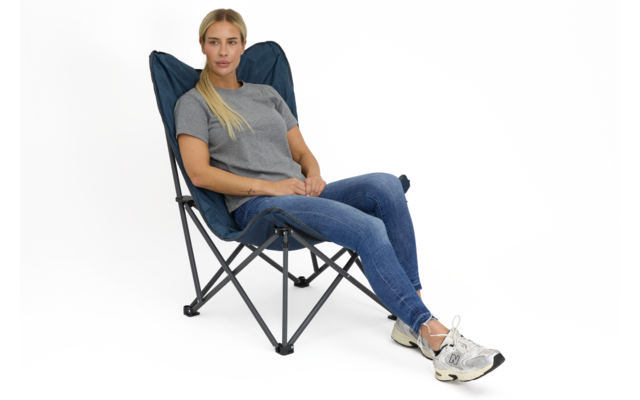 Person sitzt in blauem Klappstuhl mit schwarzem Gestell vor weißem Hintergrund, trägt graues T‑Shirt, blaue Jeans und weiße Sneakers, Hände im Schoß