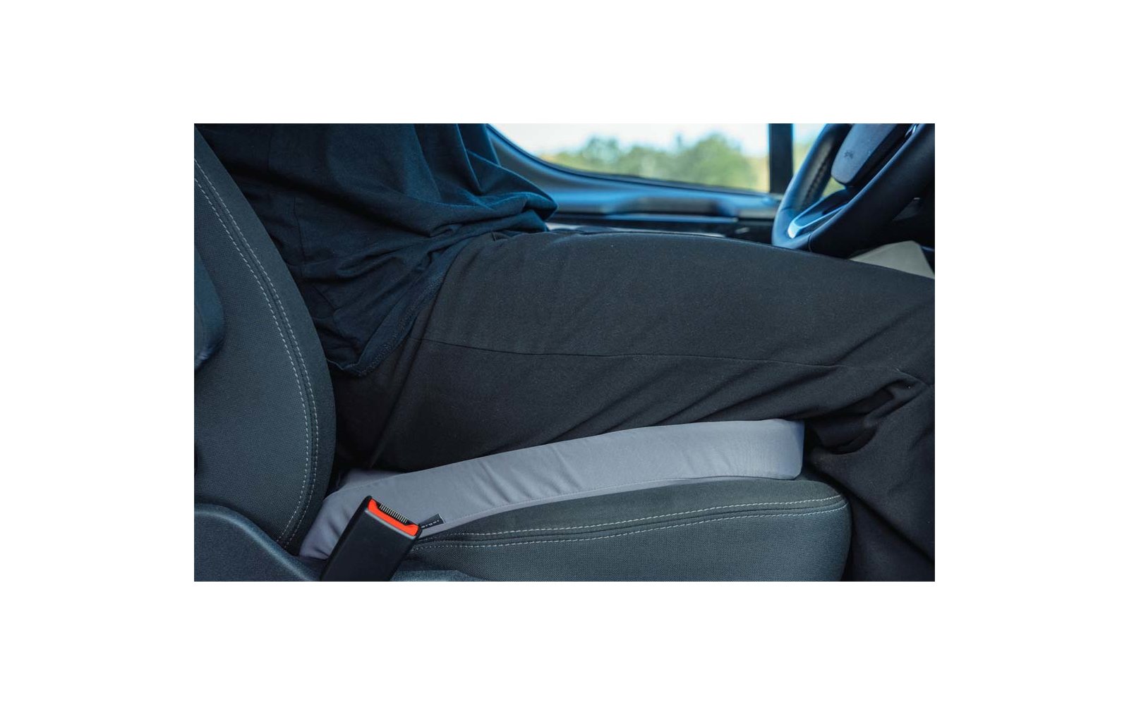 Lower body of a person wearing black clothing seated on a light gray seat cushion in a car; steering wheel and seatbelt buckle visible