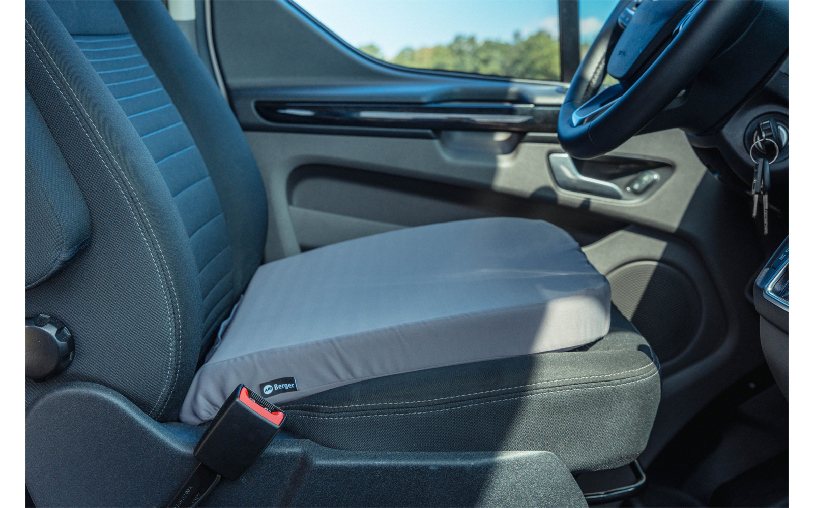 Gray seat cushion on a front car seat; visible seatbelt, door panel with handle, steering wheel and key in the ignition, outside window showing trees
