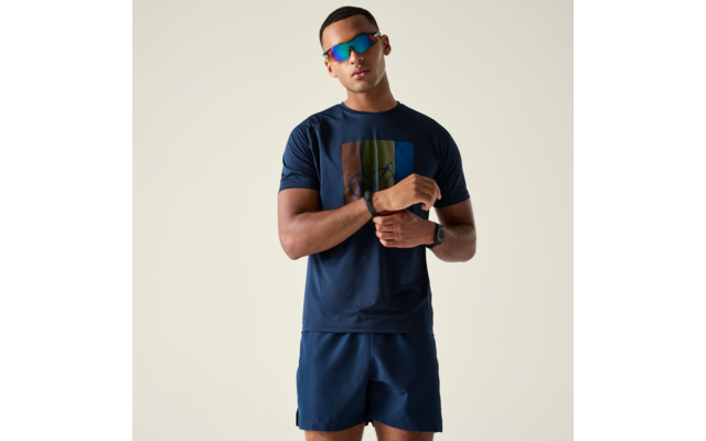 Person steht vor hellem Hintergrund, trägt dunkelblaues T‑Shirt mit farbigem Rechteck, dunkelblaue Shorts, verspiegelte Sonnenbrille und Armbanduhr, hält ein Handgelenk