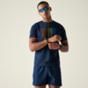 Person steht vor hellem Hintergrund, trägt dunkelblaues T‑Shirt mit farbigem Rechteck, dunkelblaue Shorts, verspiegelte Sonnenbrille und Armbanduhr, hält ein Handgelenk