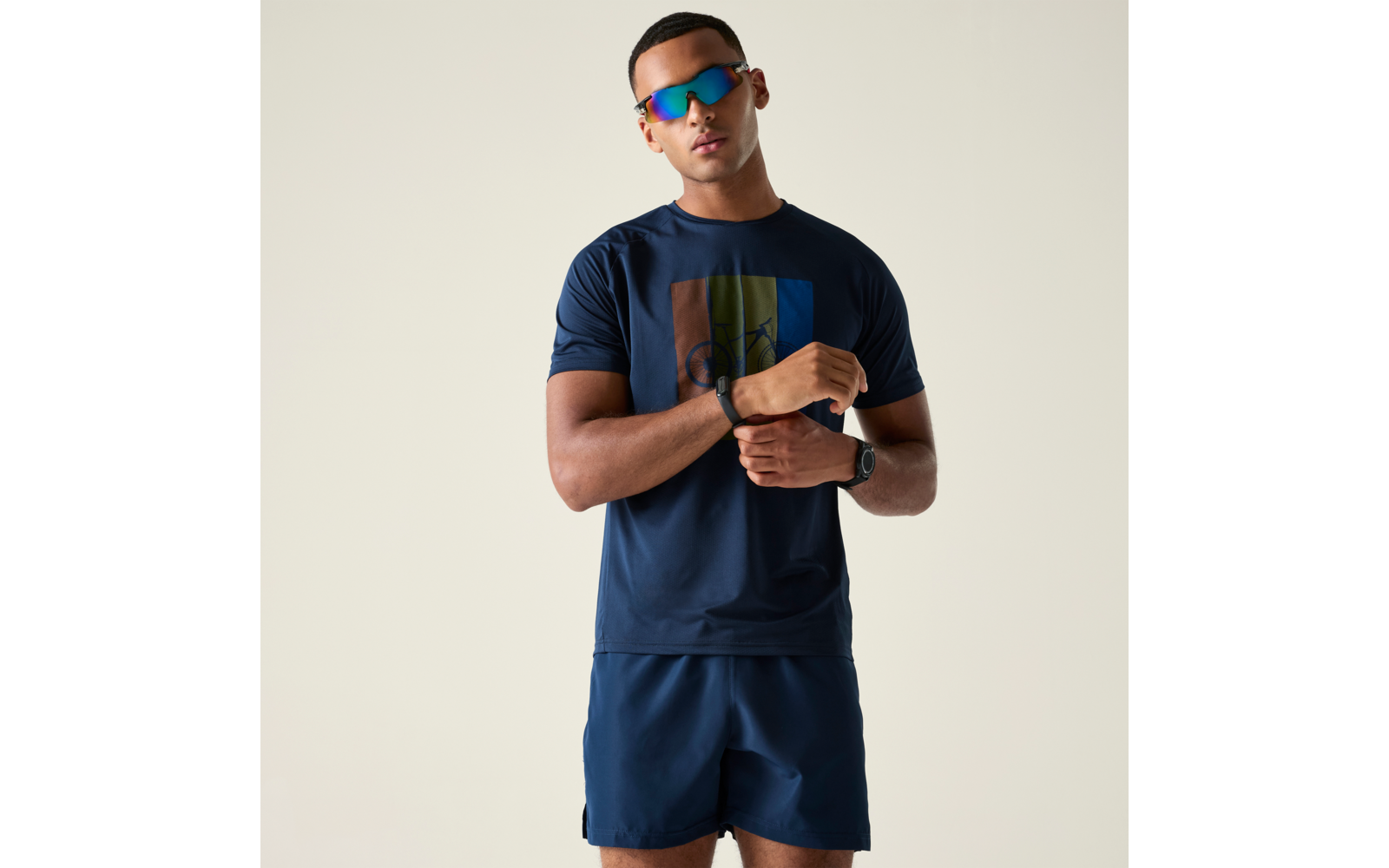 Person steht vor hellem Hintergrund, trägt dunkelblaues T‑Shirt mit farbigem Rechteck, dunkelblaue Shorts, verspiegelte Sonnenbrille und Armbanduhr, hält ein Handgelenk