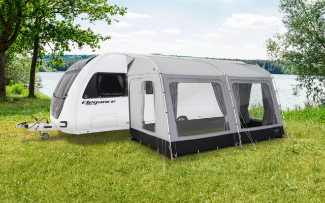 Caravan bianco con tenda annessa su un prato verde accanto a un lago e alberi