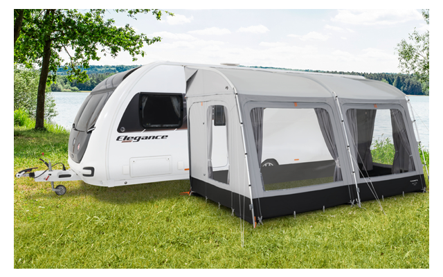 Weißer Wohnwagen mit angebautem Zelt auf einer grünen Wiese neben einem See und Bäumen