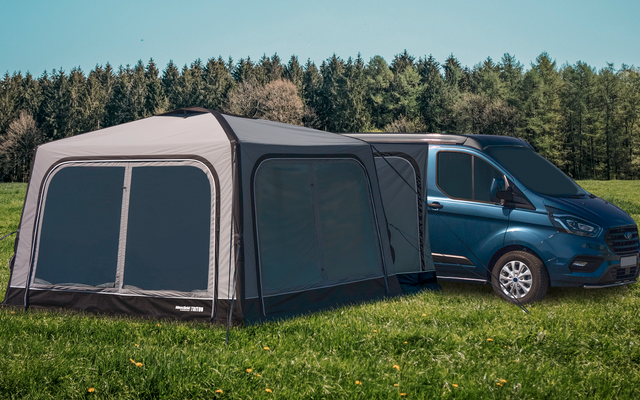 Blue van with attached gray tent on a green meadow in front of a forest