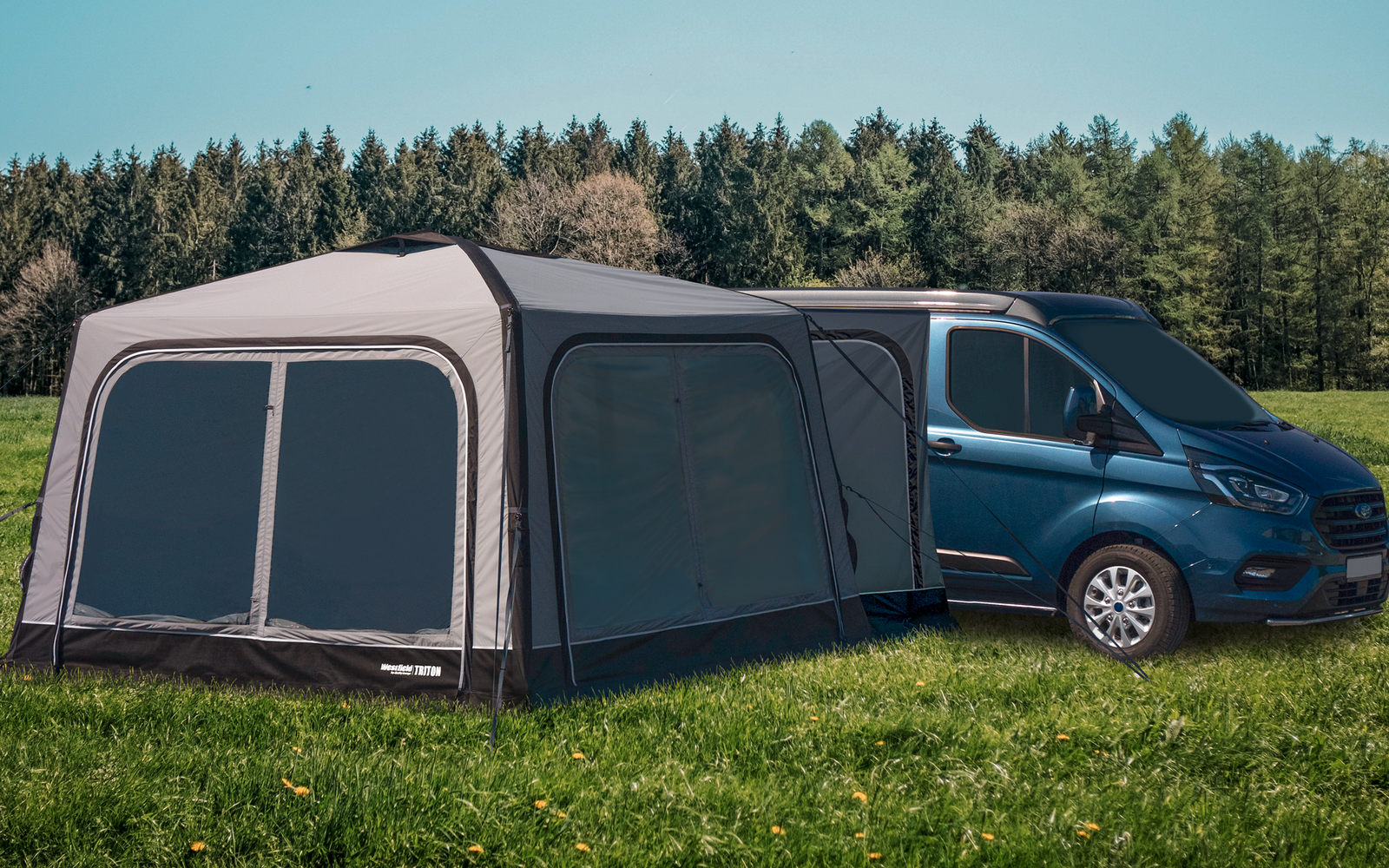 Blue van with attached gray tent on a green meadow in front of a forest