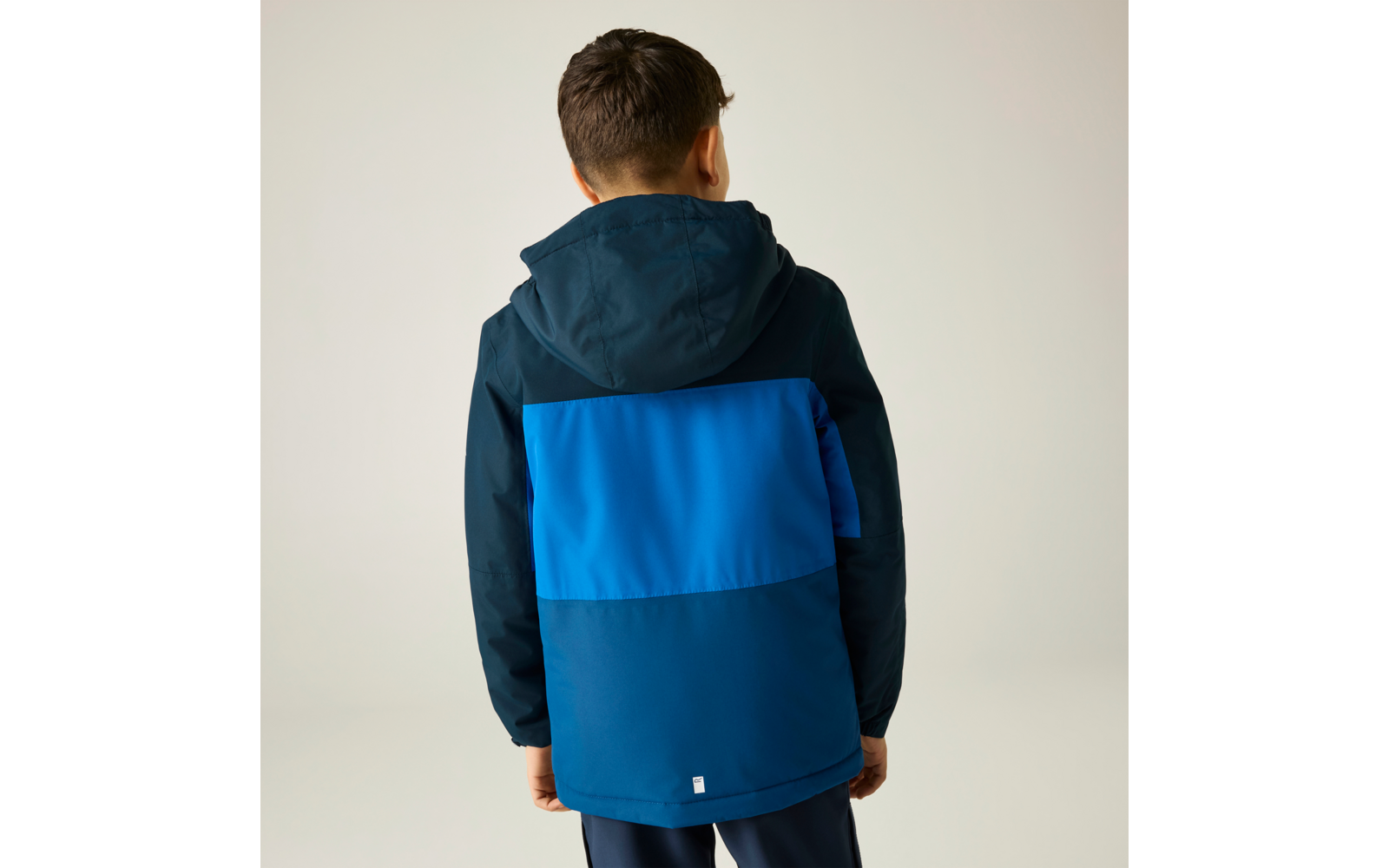 Vue de dos d'un enfant aux cheveux bruns courts portant une veste à capuche bleu bicolore et un pantalon foncé sur un fond clair