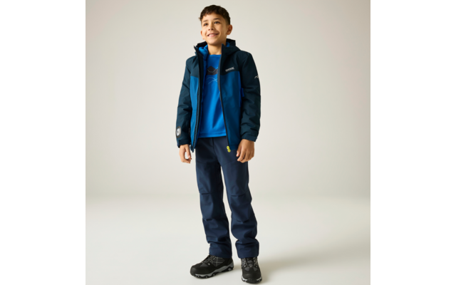 Garçon souriant debout portant une veste bleue, un sweat bleu, un pantalon bleu foncé et des chaussures de randonnée noires, fond clair