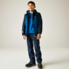 Garçon souriant debout portant une veste bleue, un sweat bleu, un pantalon bleu foncé et des chaussures de randonnée noires, fond clair