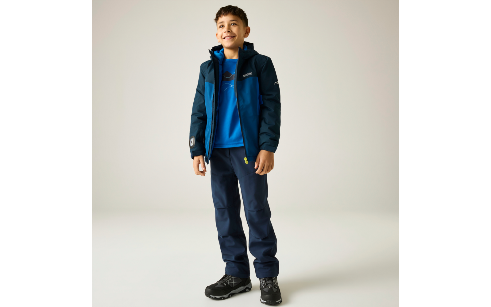 Garçon souriant debout portant une veste bleue, un sweat bleu, un pantalon bleu foncé et des chaussures de randonnée noires, fond clair