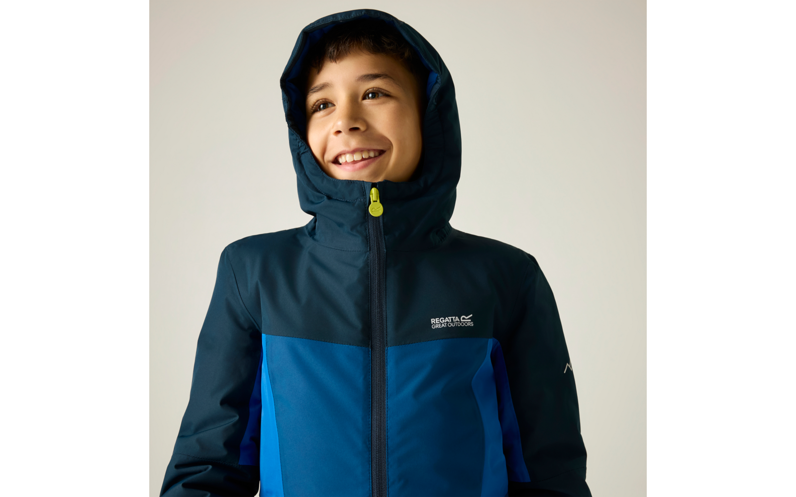 Enfant souriant portant une veste bleue à capuche, regardant vers le haut, fond clair, logo visible sur la poitrine