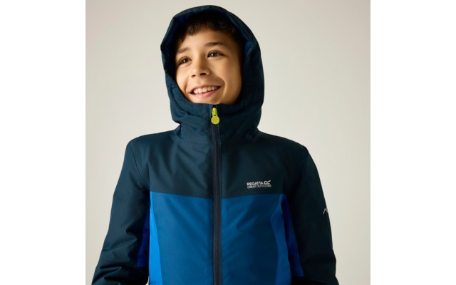 Enfant souriant portant une veste bleue à capuche, regardant vers le haut, fond clair, logo visible sur la poitrine