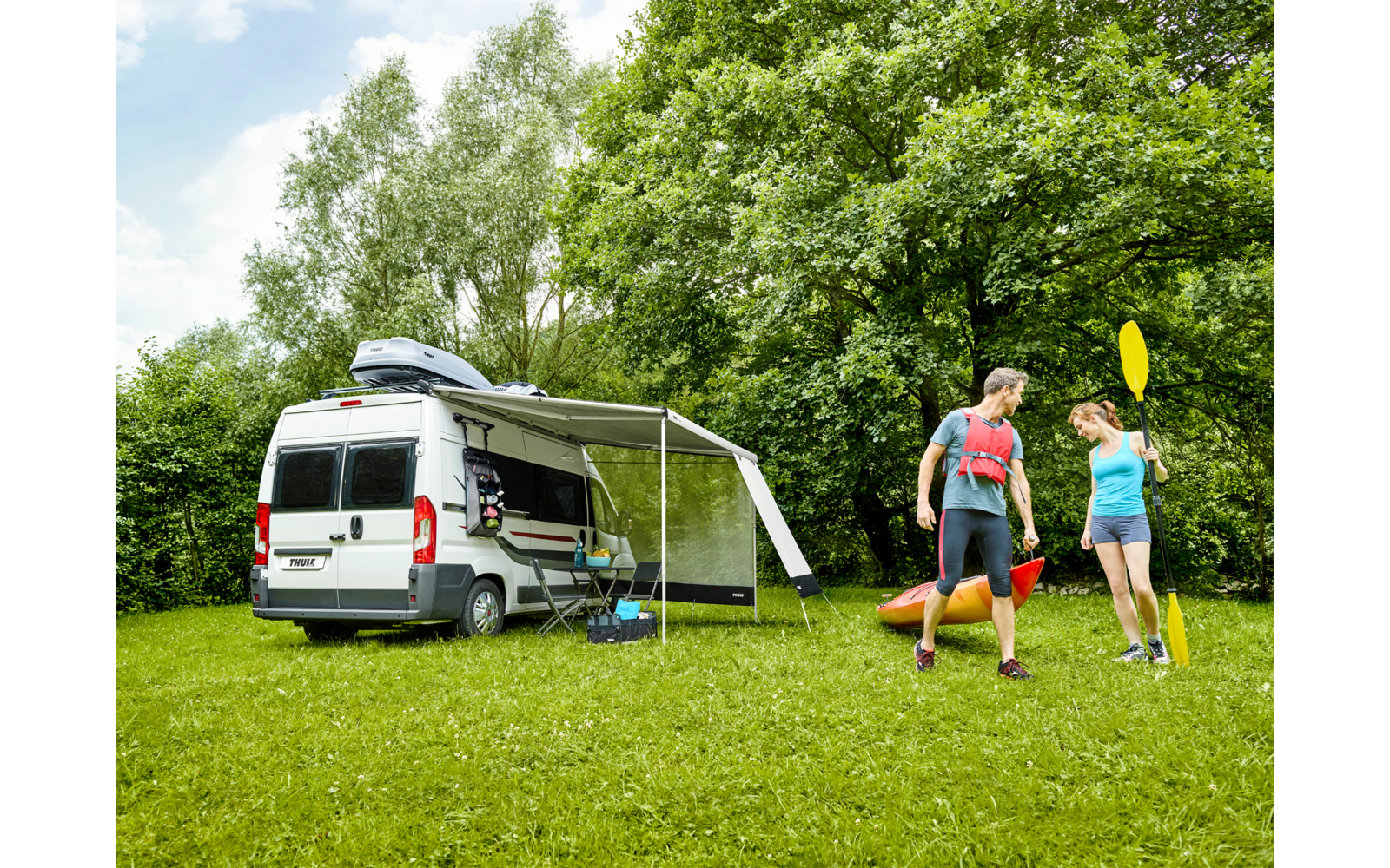 Camper bianco con tendalino aperto su un'area erbosa; due persone in piedi che tengono pagaie; due kayak colorati sull'erba; alberi a foglia e cielo azzurro sullo sfondo