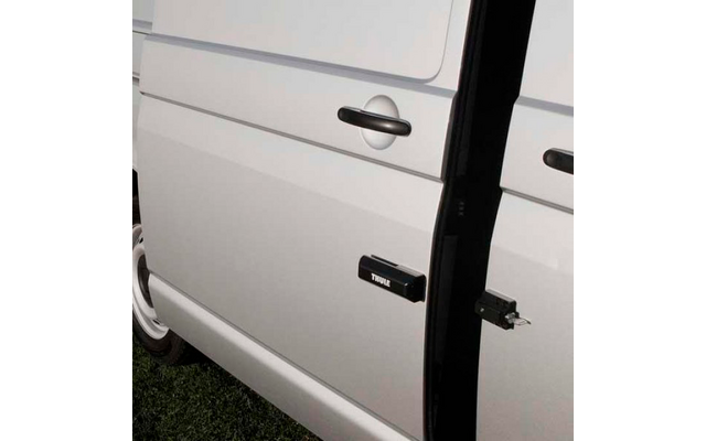 Pannello laterale bianco di un furgone con porta scorrevole parzialmente aperta, maniglia della porta, dispositivo rettangolare nero con la scritta "THULE" fissato alla porta, ruota posteriore e erba sotto
