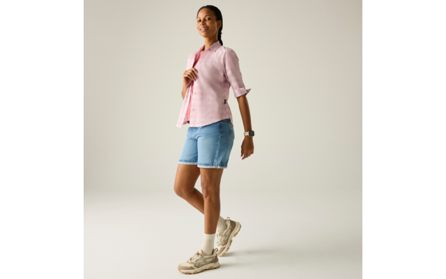 Seitlich stehende Person in rosa Bluse, Jeansshorts, Sneakers und mit Uhr vor hellem Hintergrund