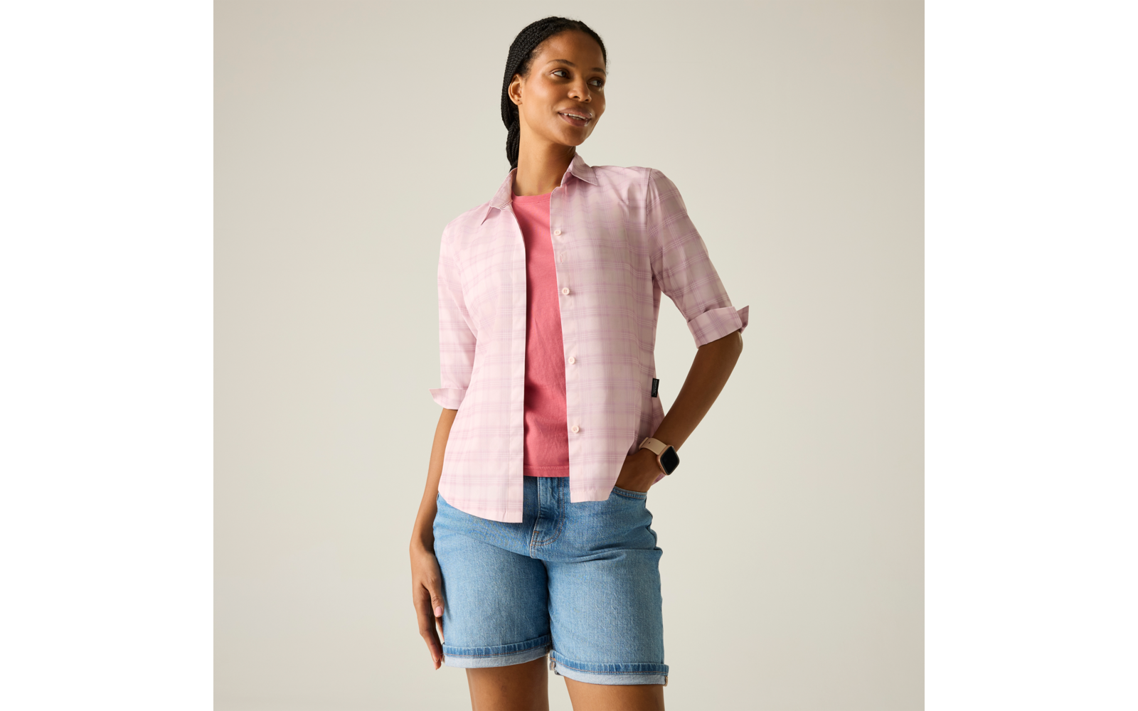 Frau in rosa kariertem Hemd über pinkem T-Shirt, Jeans-Shorts, Smartwatch; Hände in den Taschen, Blick nach rechts, heller Hintergrund