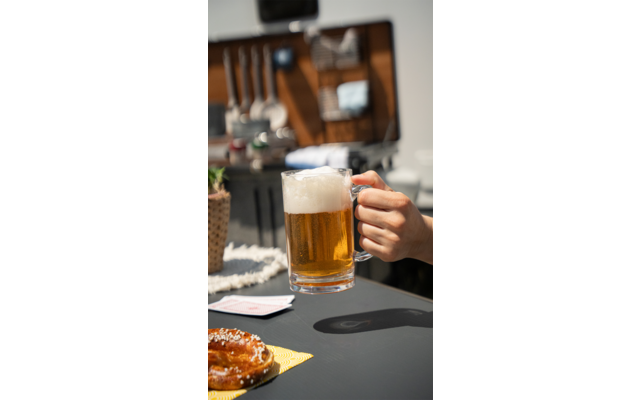 Hand hält ein Glas Bier in einem hellen Café, Tische mit Speisen und Getränken im Hintergrund