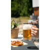 Ein Glas Bier wird von einer Hand gehalten, im Vordergrund liegt ein Teller mit Brezeln, im Hintergrund sind Pflanzen und ein Holztisch sichtbar