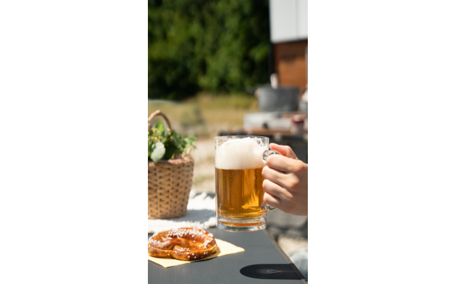 Ein Glas Bier wird von einer Hand gehalten, im Vordergrund liegt ein Teller mit Brezeln, im Hintergrund sind Pflanzen und ein Holztisch sichtbar