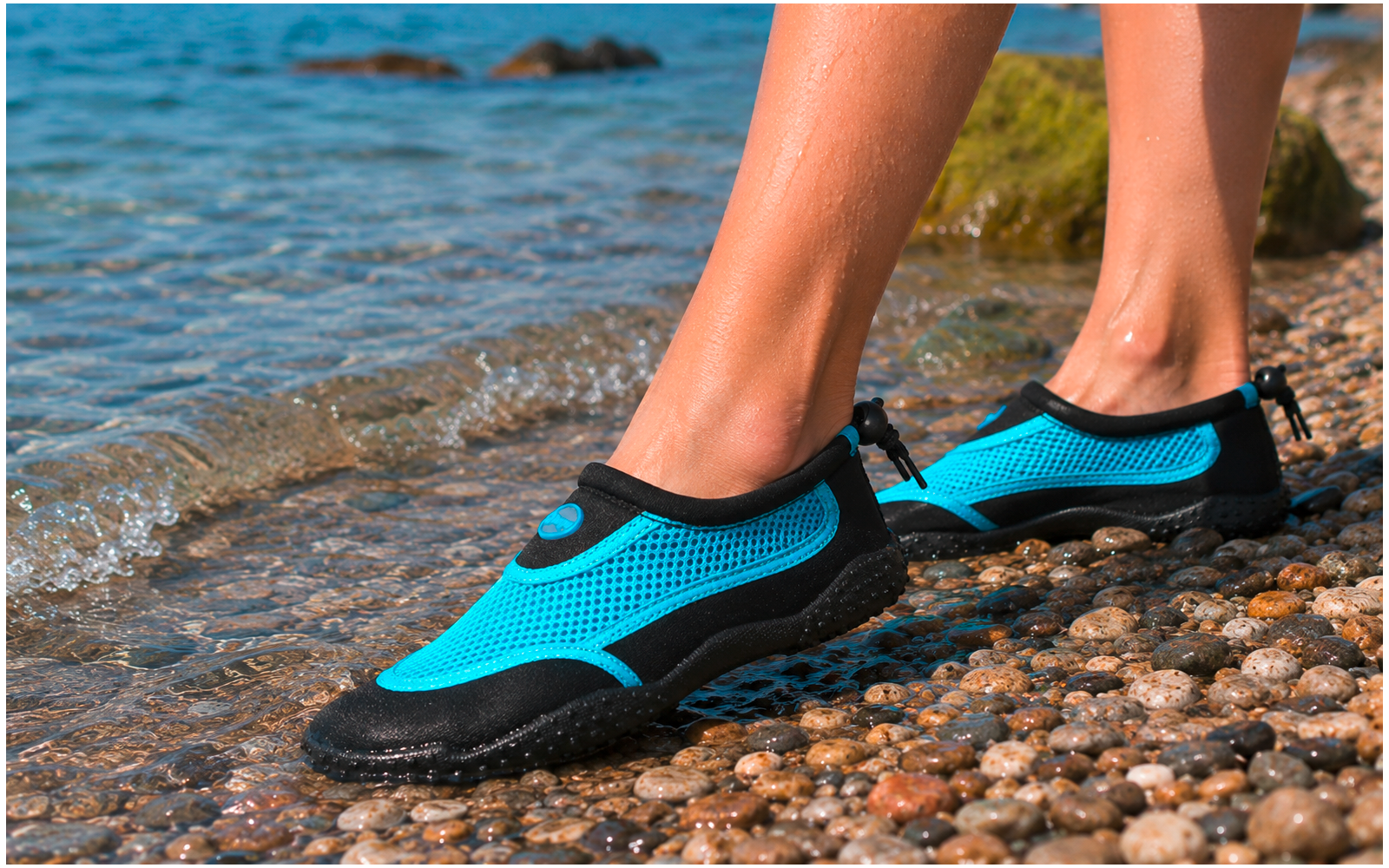 Piernas inferiores y pies con zapatos de agua azules sobre una orilla de guijarros, agua poco profunda con pequeñas olas y una roca cubierta de musgo al fondo