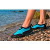Piernas inferiores y pies con zapatos de agua azules sobre una orilla de guijarros, agua poco profunda con pequeñas olas y una roca cubierta de musgo al fondo