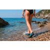 Persona vista de espaldas caminando en la orilla de una playa de guijarros, lleva braguita de baño negra y zapatillas de agua turquesas; rocas y mar visibles