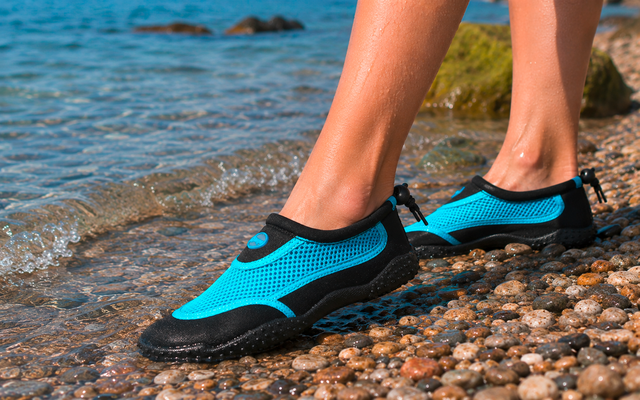 Piernas inferiores y pies con zapatos de agua azules sobre una orilla de guijarros, agua poco profunda con pequeñas olas y una roca cubierta de musgo al fondo