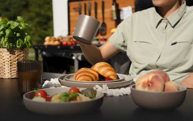 Torso einer Person in hellgrünem Hemd hält eine Metalltasse über einem Teller mit zwei Croissants; auf dem Tisch Schalen mit Salat und Pfirsichen, ein Glas mit Getränk, ein geflochtener Korb mit Kräuterpflanze; im Hintergrund unscharfes Holzbrett mit