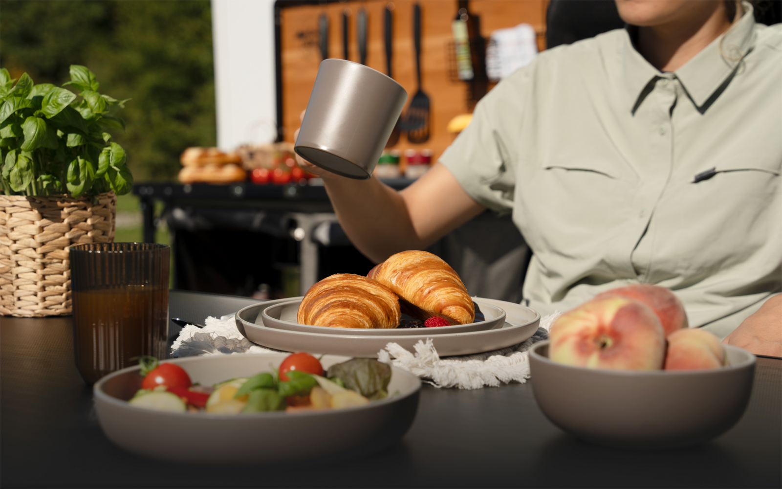 Torso einer Person in hellgrünem Hemd hält eine Metalltasse über einem Teller mit zwei Croissants; auf dem Tisch Schalen mit Salat und Pfirsichen, ein Glas mit Getränk, ein geflochtener Korb mit Kräuterpflanze; im Hintergrund unscharfes Holzbrett mit