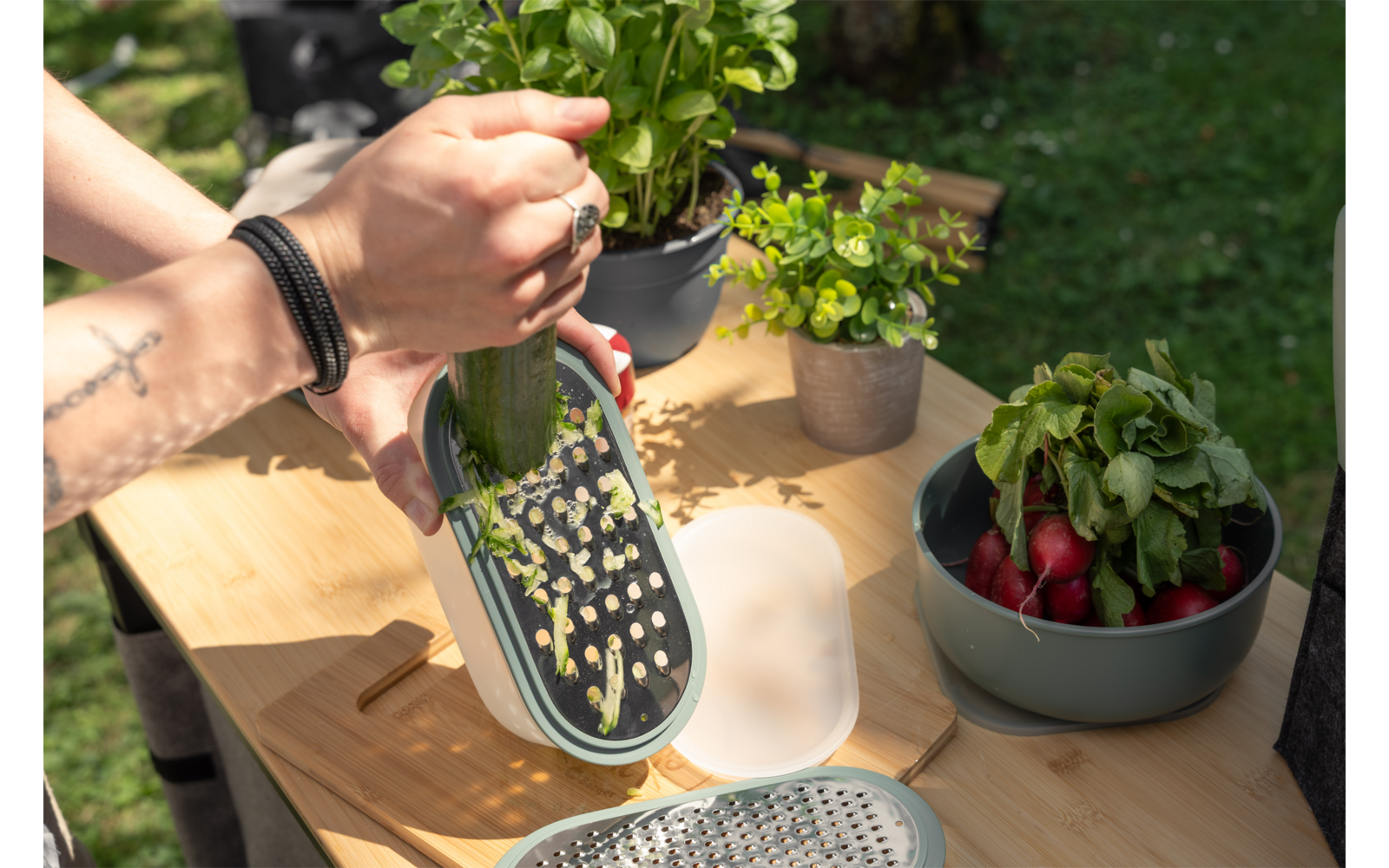Handen raspen een komkommer met een rasp boven een bakje op een houten tafel; zichtbaar een getatoeëerde arm met armband, bloempotten en een kom radijsjes