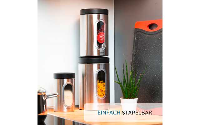 Three silver cylindrical canisters with black lids and viewing windows on a wooden shelf; windows show red round pieces, short yellow tubes, and white fine-grained material; beside them a small green potted plant and a black cutting board with a red edge