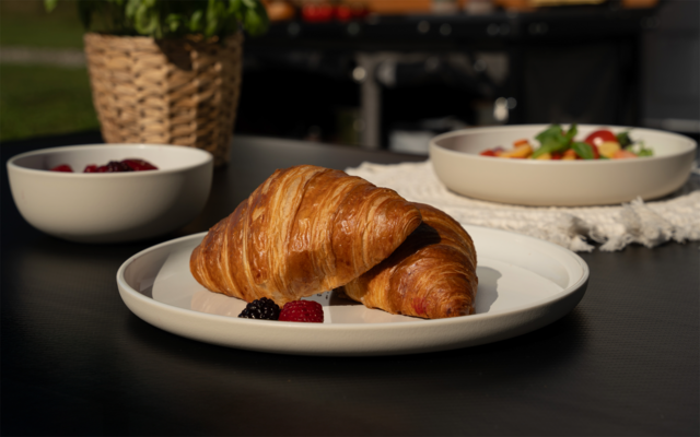Zwei Croissants auf weißem Teller mit einer Brombeere und einer Himbeere; im Hintergrund zwei weiße Schalen, ein geflochtener Korb mit Pflanze und ein Grill