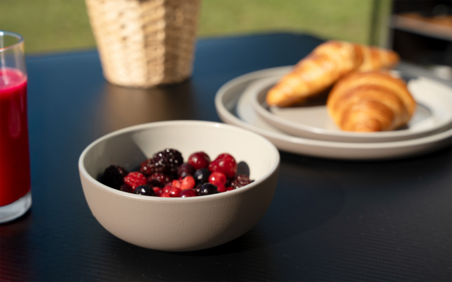Bol de fruits rouges mélangés sur une table foncée ; à l'arrière-plan une assiette avec deux croissants, un verre de jus rouge et un panier tressé