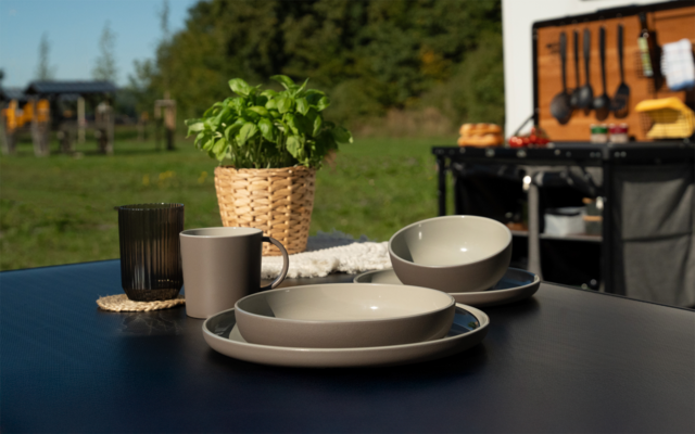 Table extérieure noire avec deux assiettes, deux bols, deux tasses, un verre et un basilic dans un pot tressé ; en arrière-plan une cuisine de camping avec ustensiles suspendus et prairie