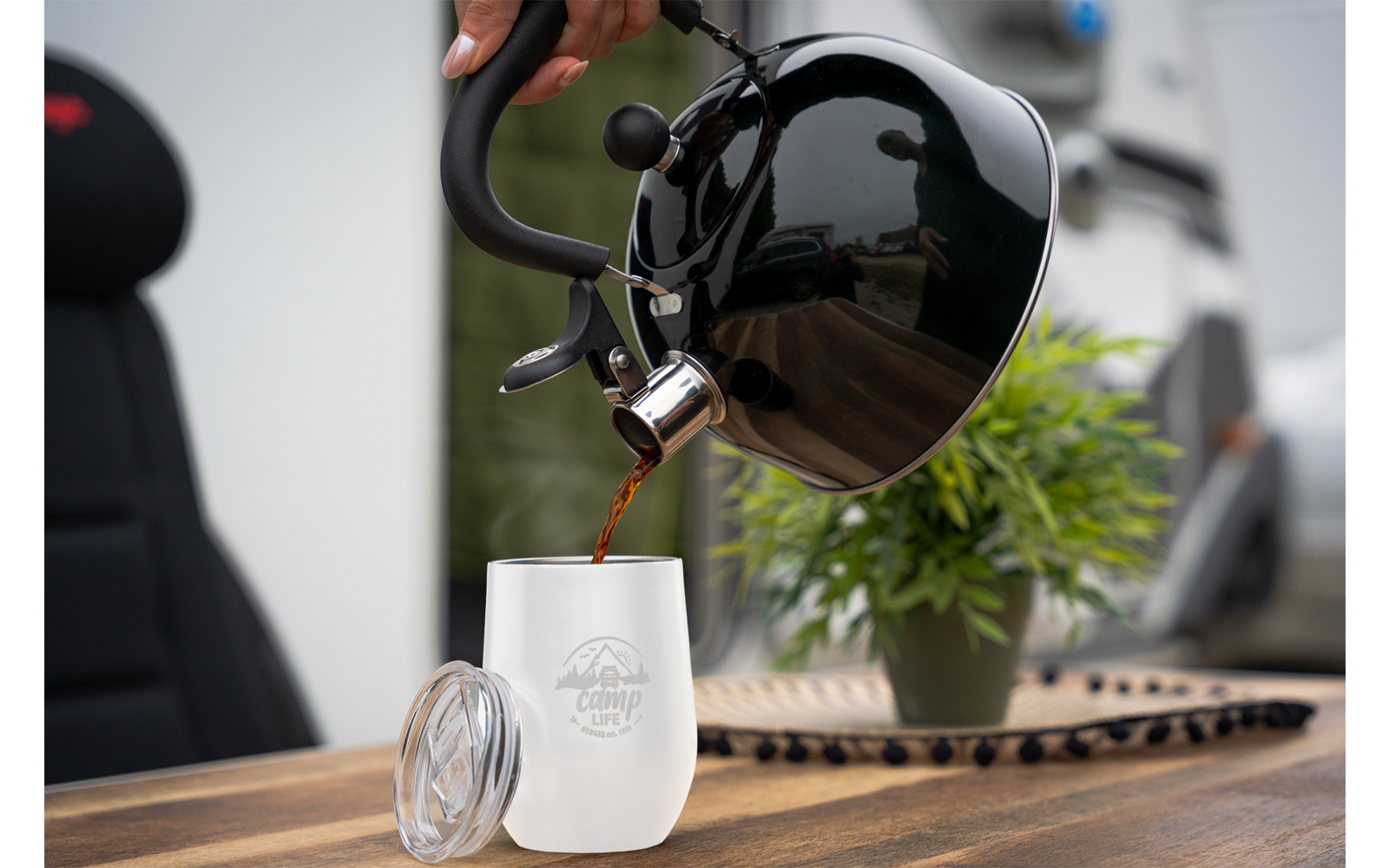 Zwarte waterkoker giet heet water in een wit glas met deksel, plant en stoel op houten tafel op de achtergrond