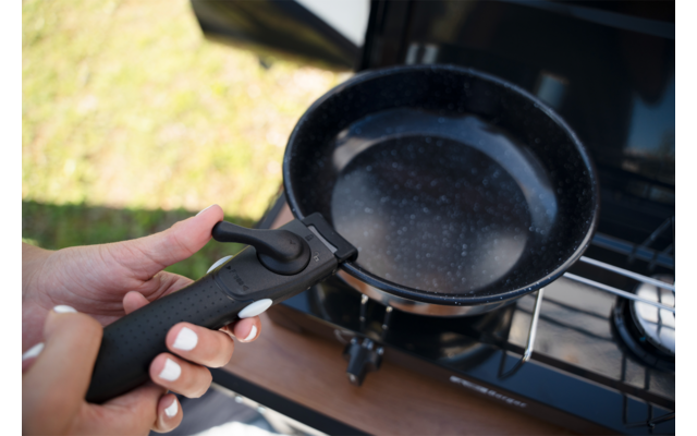 Zwei Hände halten einen abnehmbaren schwarzen Pfannengriff über einer schwarzen Pfanne mit hellen Sprenkeln auf einem Gaskocher; unscharfes Gras im Hintergrund
