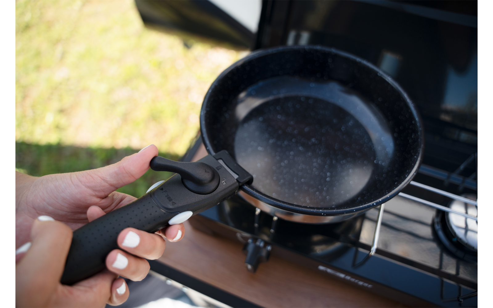 Zwei Hände halten einen abnehmbaren schwarzen Pfannengriff über einer schwarzen Pfanne mit hellen Sprenkeln auf einem Gaskocher; unscharfes Gras im Hintergrund
