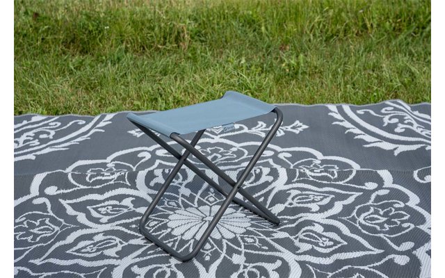 Opvouwkrukje met blauw stoffen zitje en zwarte gekruiste poten op een grijs-wit patroonkleed; gras op de achtergrond