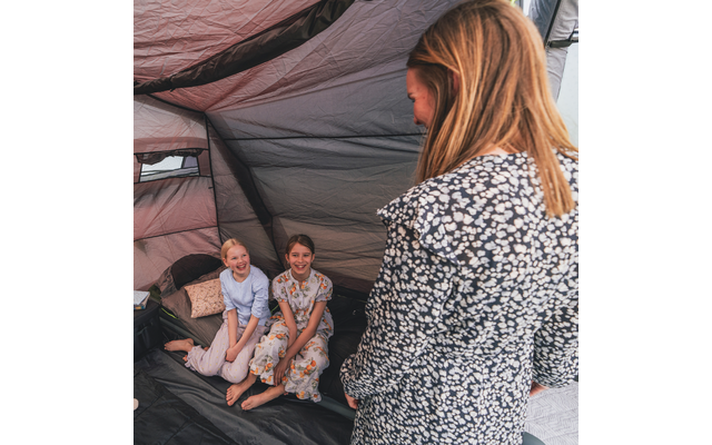 Zwei lächelnde Kinder sitzen barfuß nebeneinander auf dem Boden eines Zelts. Eine Person mit floralem Oberteil steht mit dem Rücken zur Kamera. Zeltinnenraum mit Kissen und Gepäck sichtbar