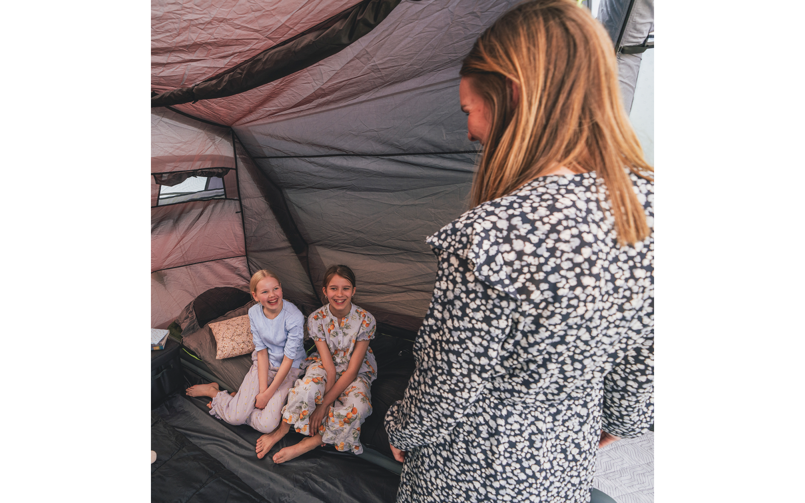 Zwei lächelnde Kinder sitzen barfuß nebeneinander auf dem Boden eines Zelts. Eine Person mit floralem Oberteil steht mit dem Rücken zur Kamera. Zeltinnenraum mit Kissen und Gepäck sichtbar