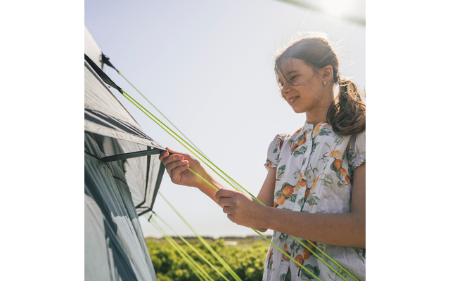 Eine Person hält grüne Zeltleinen neben einem Zelt; im Hintergrund Gebüsch und heller Himmel