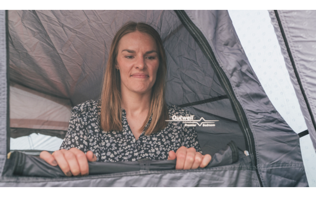 Frau mit schulterlangen braunen Haaren und gemusterter Bluse hält den Rand einer Zelttür mit sichtbarem Reißverschluss, Zeltinnenraum mit Logo sichtbar