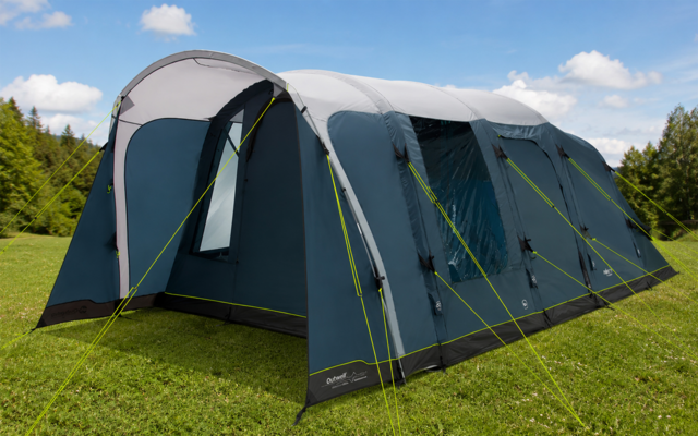 Groot blauw-grijs tunneltent met meerdere bogen en neongroene scheerlijnen op een grasveld, bomen op de achtergrond, blauwe lucht met enkele wolken