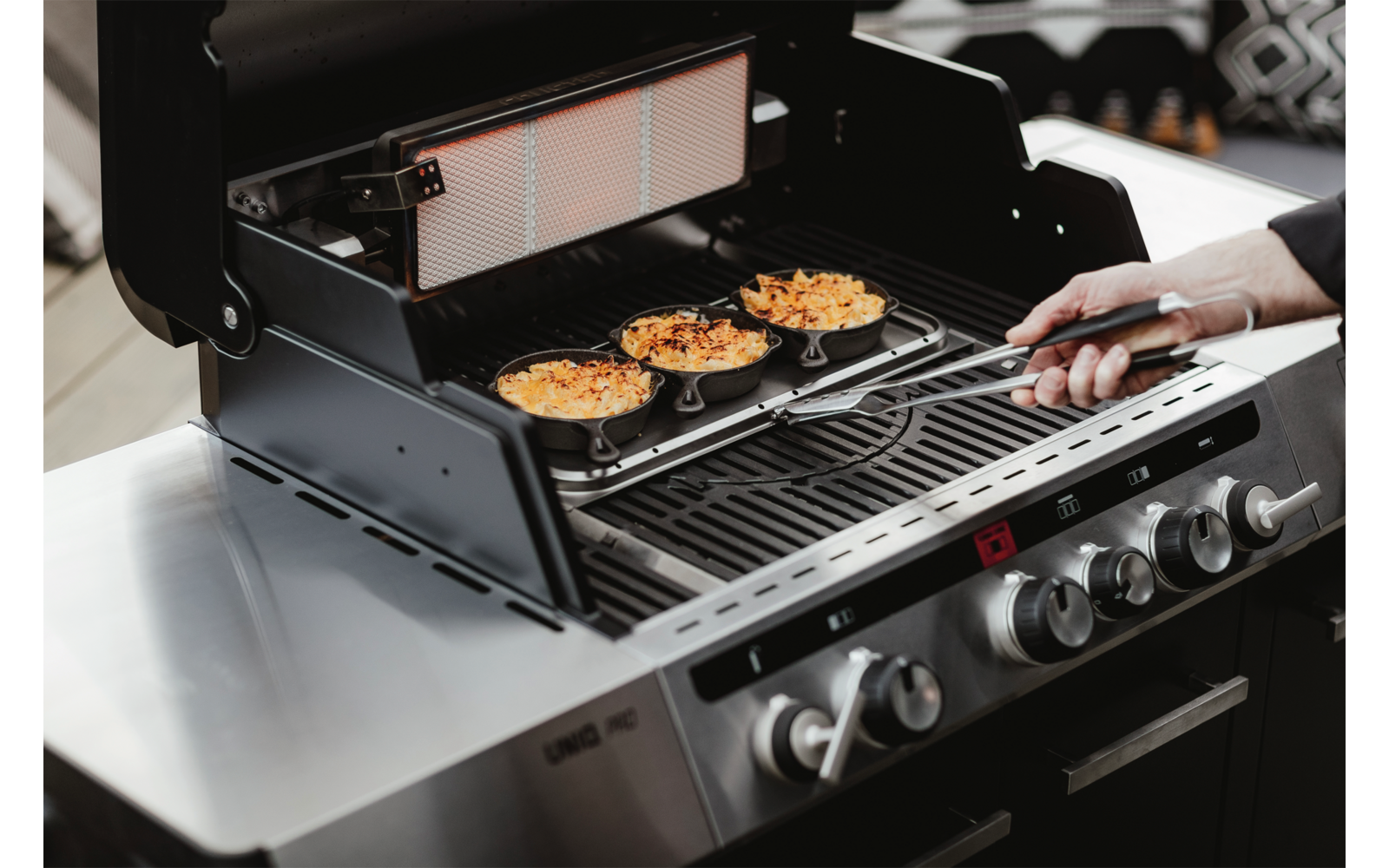 Ein Gasgrill mit mehreren Burger-Patties auf dem Rost und einer Hand, die eine Grillzange hält