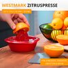 Main presse une orange coupée sur une presse à agrumes rouge sur un plan de travail en bois; orange coupée à côté, corbeille métallique avec oranges et citrons verts; mention "WESTMARK ZITRUSPRESSE" en haut