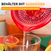 Récipient plastique rouge avec bec verseur versant un liquide rose-orangé dans un verre ; pamplemousse coupé en deux et tranches de fruits au premier plan, main tenant le récipient